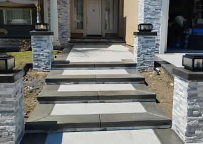 Concrete steps with black trim lead to a beige house entrance, flanked by stone pillars with modern lanterns and a small palm tree on the left.